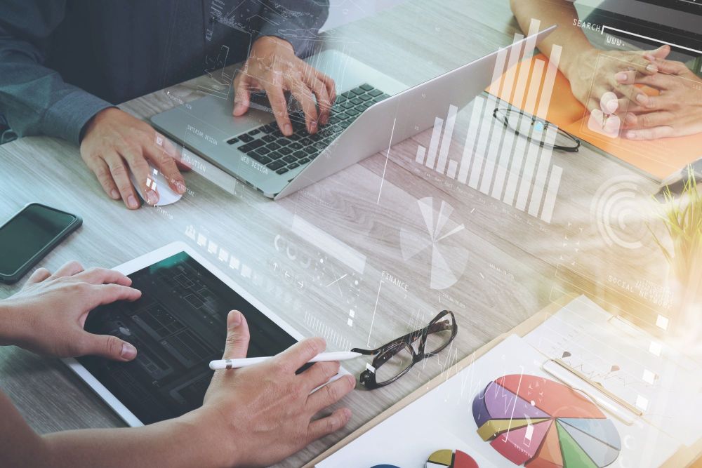 business documents on office table with smart phone and laptop computer and graph business with social network diagram and three colleagues discussing data in the background